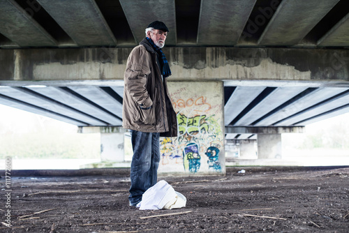 Lost homeless man standing with bag under bridge.