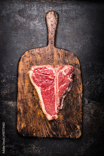 Raw T-bone steak on aged wooden cutting board on dark rust metal background, top view, close up photo