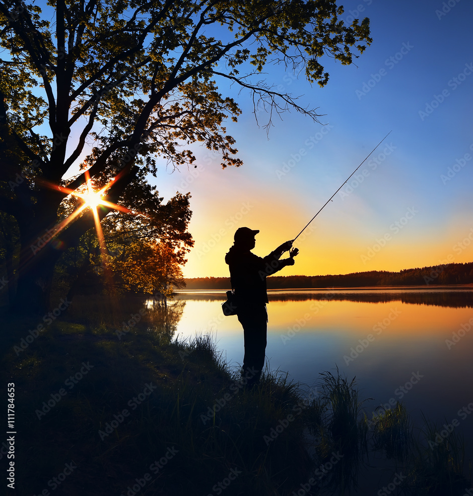 silhouette of a fisherman with a fishing rod on the shore of the lake, the  river in the morning. The rays of the rising sun filtering through the tree  leaves Stock Photo