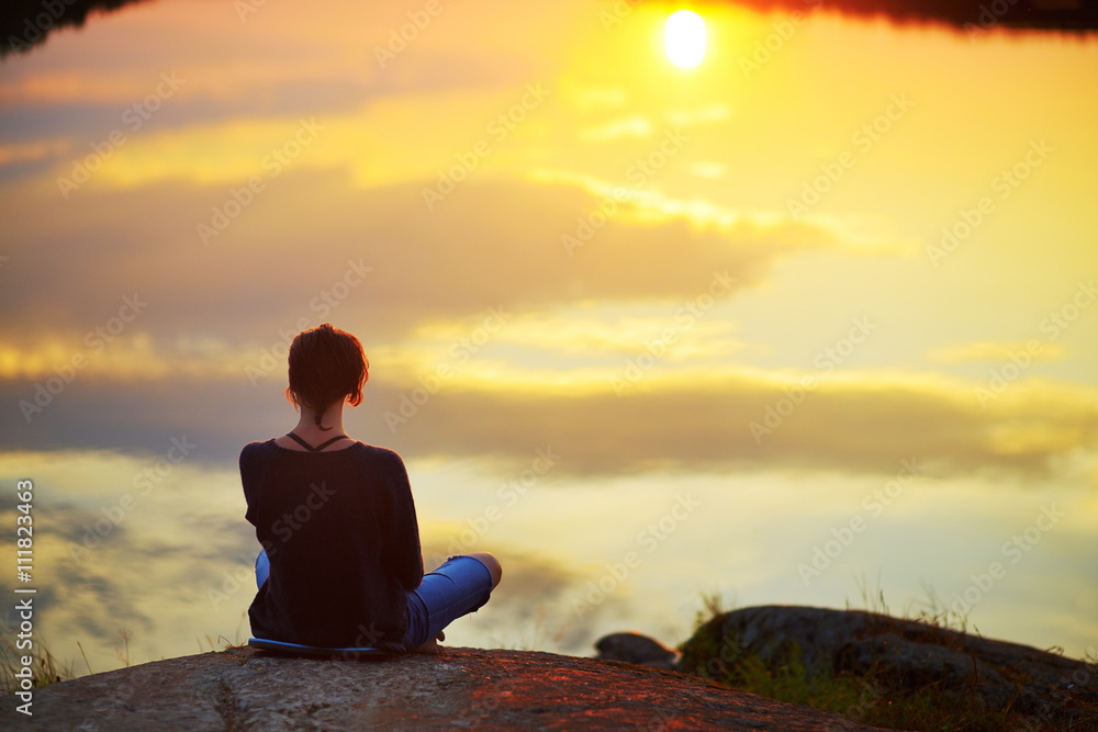 Young woman sitting on the stone enjoying peaceful moment of sunset. In the reflection of the lake water sees clouds and sun.