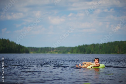 relaxed young man lying on inflatable ring in lake and admiring the stunning views. Away on a blurred background forest and sky.
