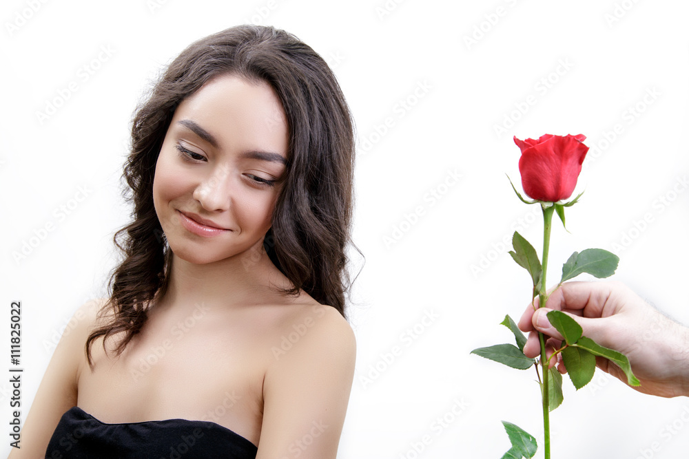 Beautiful angry girl receives one red rose. She is surprised, looking at the flowers and starts smiling. Men's hand holding one rose. Girl is white with bushy brown hair. Isolated on white background.