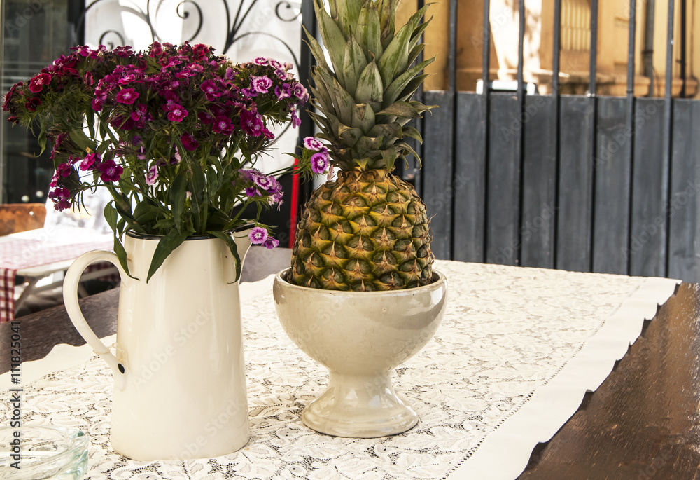 Fototapeta premium Jug with flowers and pineapple on table of street cafe