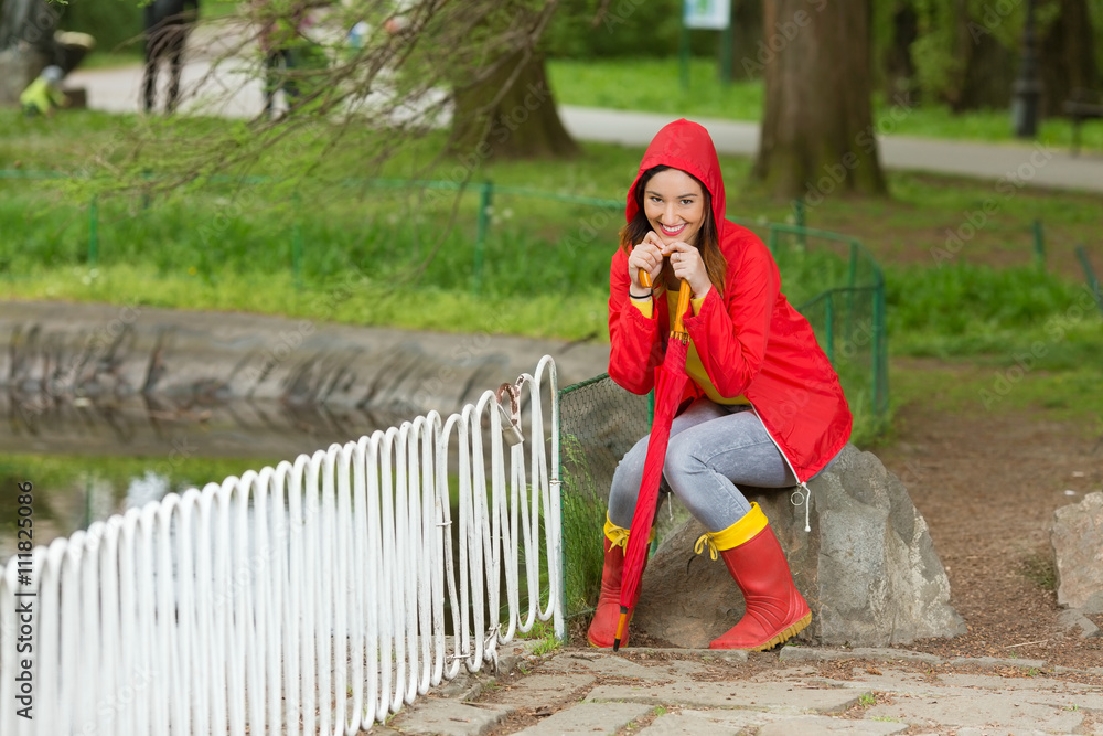 Beautiful young woman is sitting on the stone near the pond in a park on a rainy spring day. She is wearing red raincoat and rubber boots and leaning on a red umbrella. Beautiful young woman is sitting on the stone near the pond in a park on a rainy spring day. She is wearing red raincoat and rubber boots and leaning on a red umbrella.