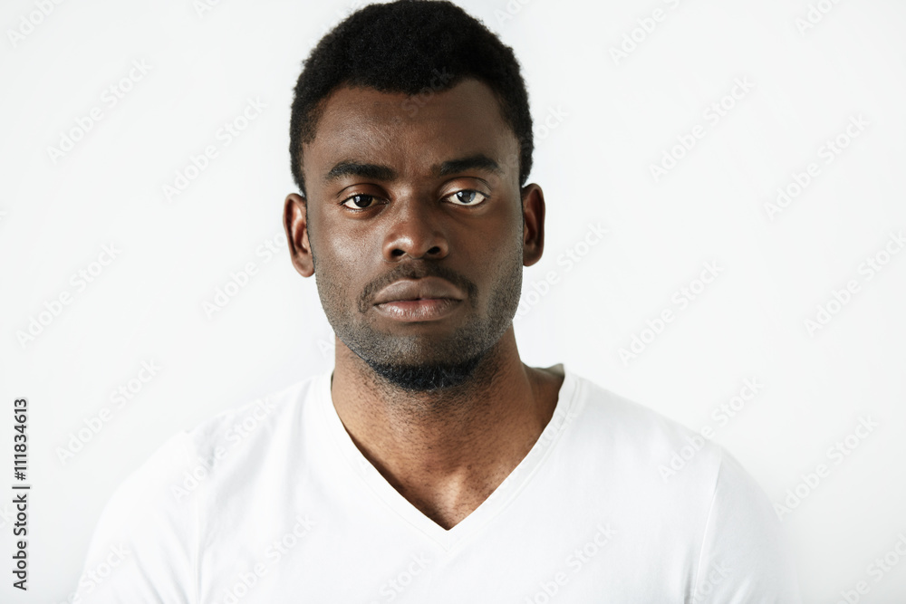 Close up isolated view of serious handsome young African male in white T-shirt looking with thoughtful expression at the camera. Lifestyle and people concept. Human expressions, emotions, feelings Close up isolated view of serious handsome young African male in white T-shirt looking with thoughtful expression at the camera. Lifestyle and people concept. Human expressions, emotions, feelings