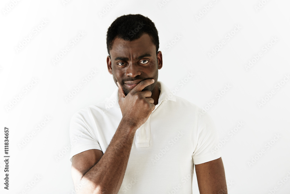 Skeptical young black man looking at the camera with suspicious or annoyed expression. Cautious and thoughtful African male thinking with hands on his chin. Human emotions and facial expressions Skeptical young black man looking at the camera with suspicious or annoyed expression. Cautious and thoughtful African male thinking with hands on his chin. Human emotions and facial expressions