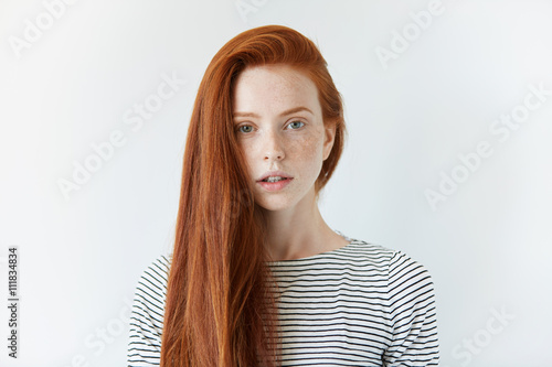Pretty student girl with perfect healthy freckled skin looking at the camera with serious expression. Redhead model with green eyes wearing striped top standing against white studio wall background