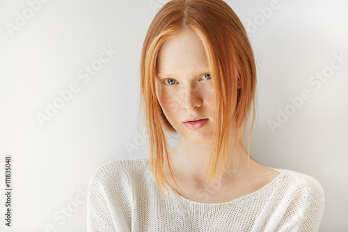 Close up portrait of beautiful freckled Caucasian teenage girl with red hair and blue eyes looking and smiling at the camera. Student girl posing against white copy space studio wall background