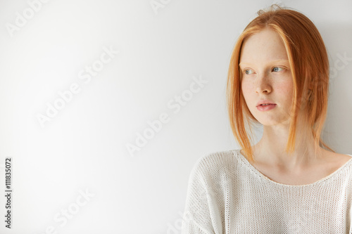 Isolated headshot of pretty teenage girl with ginger hair and perfect freckled skin holding arms folded. Cute young female wearing stylish white top  looking away at the blank copy space wall