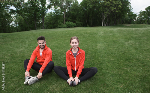 Sport man and woman sitting on green grass in park or forest. Happy couple looking at camera and smiling. Meditation and yoga concepts.