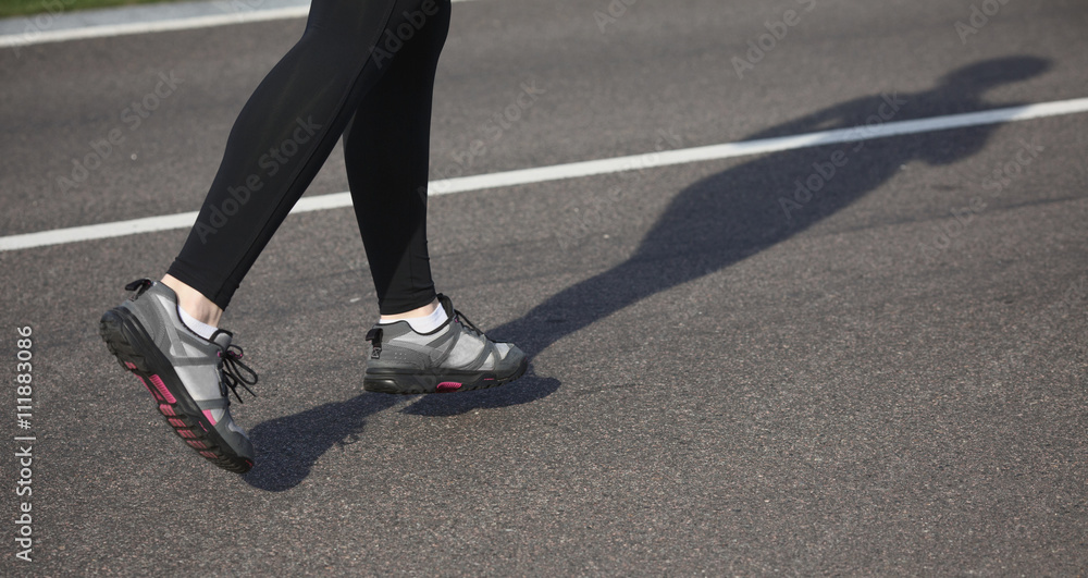 Jogging woman on track. Legs of woman running on road near park or forest. Closeup of female in running shoes going for run on road at sunrise or sunset.