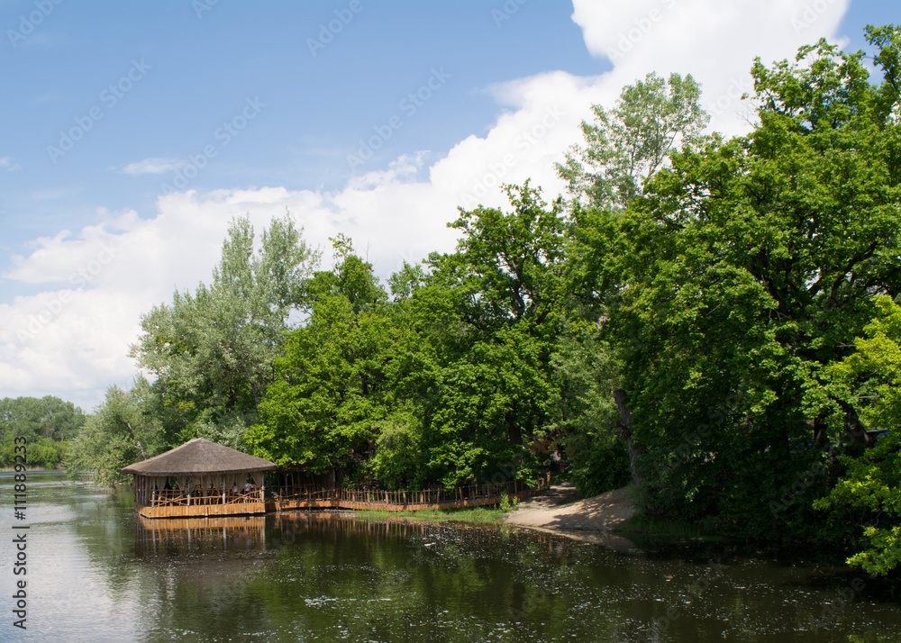 River bank: spring landscape. Blue sky, green trees and small wooden summerhouse on the water River bank: spring landscape. Blue sky, green trees and small wooden summerhouse on the water