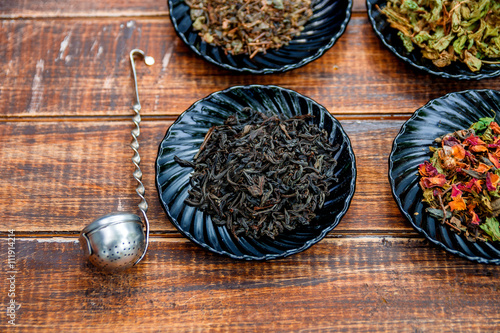 Different kinds of tea on plates on wooden background near vintage strainer. Assortment of dry tea. Tea concept. Tea leaves. Top view. Closeup