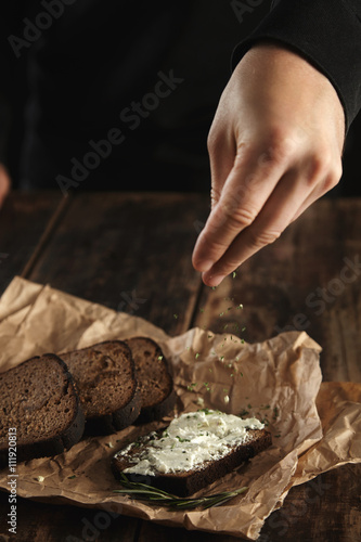 Hand pours some herb spicies on sandwich with rustic bread and soft goat cheese with herbs near slices of homemade luxury rye bread with figs and rosemary spice on craft paper on wooden table
