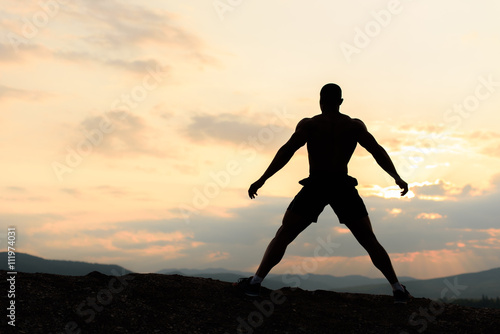 Silhouette of bodybuilder posing at the sunrise or sunset in mountains. Handsome strong man showing his muscles