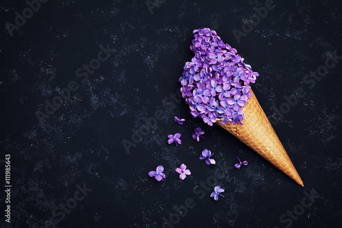 Fototapeta Naklejka Na Ścianę i Meble -  Ice cream of lilac flowers in waffle cone on black background from above, beautiful floral arrangement, vintage color, flat lay styling