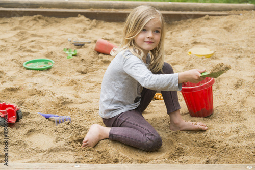 Billede på lærred Little blonde girl is playing in sandbox with plastic toy tools