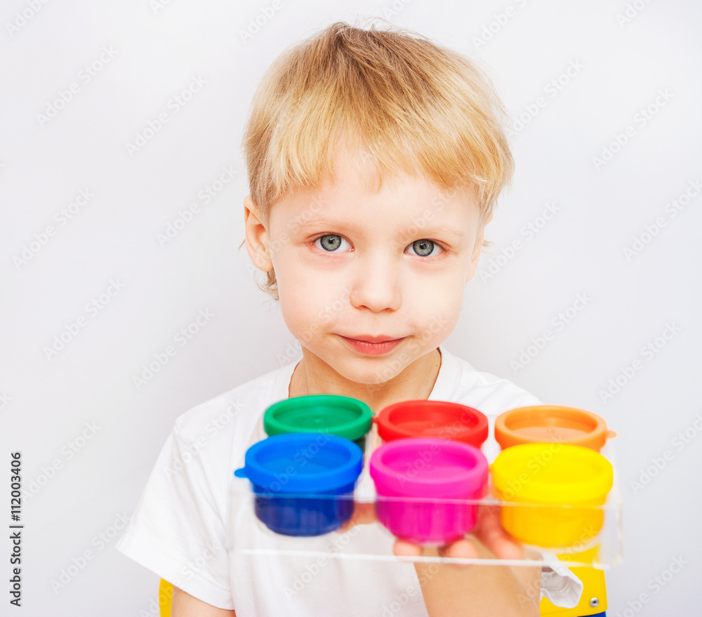 Beautiful baby boy holding new set of colored finger paints for children looking at camera. Bright colored cans and child isolated on white background. Red, blue, pink, orange, green, yellow colors Beautiful baby boy holding new set of colored finger paints for children looking at camera. Bright colored cans and child isolated on white background. Red, blue, pink, orange, green, yellow colors