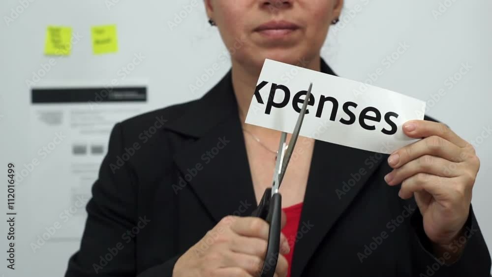 Female office worker or business woman cuts a piece of paper with the word expenses on it as an expense reduction business concept. Female office worker or business woman cuts a piece of paper with the word expenses on it as an expense reduction business concept.