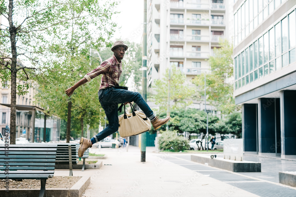 Young handsome afro black man jumping in the street of the city, looking upward, holding a bag - jumping, having fun concept
