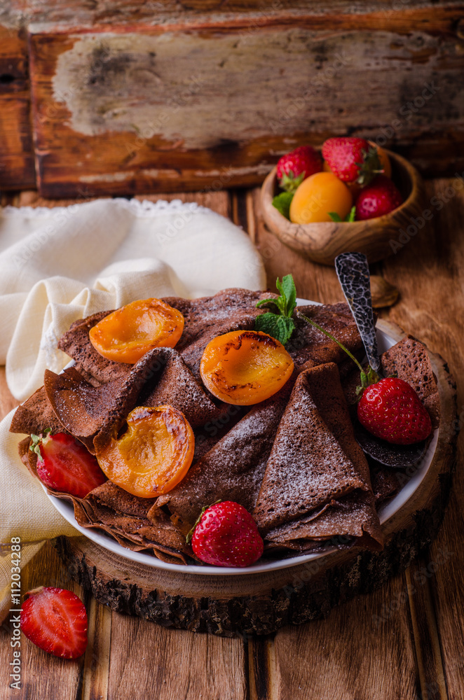 Stack of chocolate crepes decorated with strawberry and grilled apricots on wooden background. Selective focus.