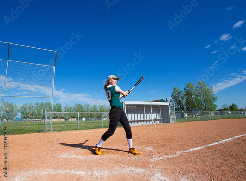A teenage girl wearing green and black baseball uniform standing at home plate on a baseball diamond swinging a bat at a baseball © kat7213