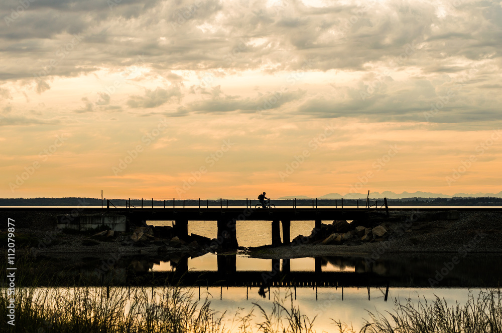 Man bicycling on a railroad trestle over a pond on a cloudy sunset evening.