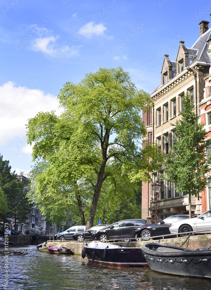 Fototapeta premium Canal of Amsterdam with green trees and boats anchored at the canal. Idyllic water street in Amsterdam, the Netherlands.