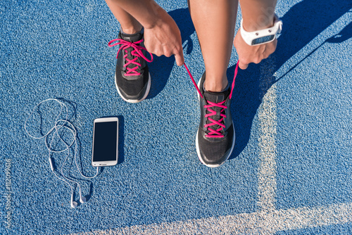 Fotografie Runner woman tying running shoes laces getting ready for race on run track with smartphone and earphones for music listening on mobile phone