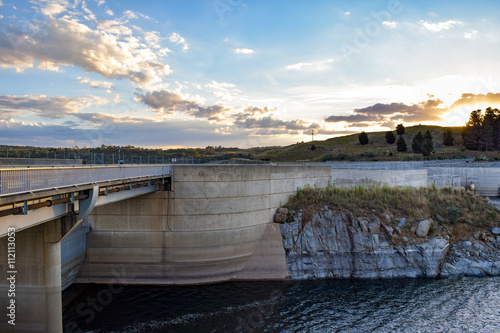 Kosciuszko Road running over the Jindabyne Dam wall with mountain view background and cloudscape.  Lake Jindabyne is part of the ground breaking 'Snowy Mountains Hydro-Electric Scheme'.