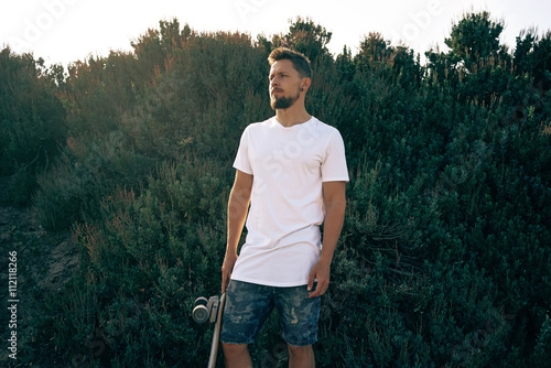 A male wearing a blank white t-shirt and shorts is standing with a skateboard on a green background. photo