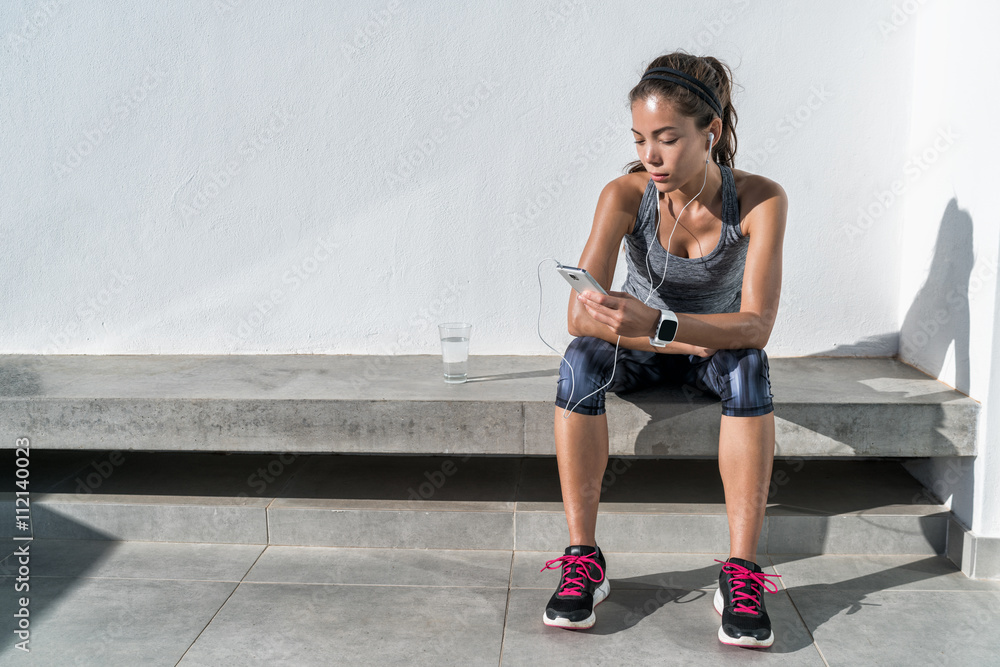 Fototapeta premium Young woman listening to music with earphones on smart phone app for fitness motivation. Athlete runner in sportswear relaxing sitting getting inspired. Asian mixed race model.