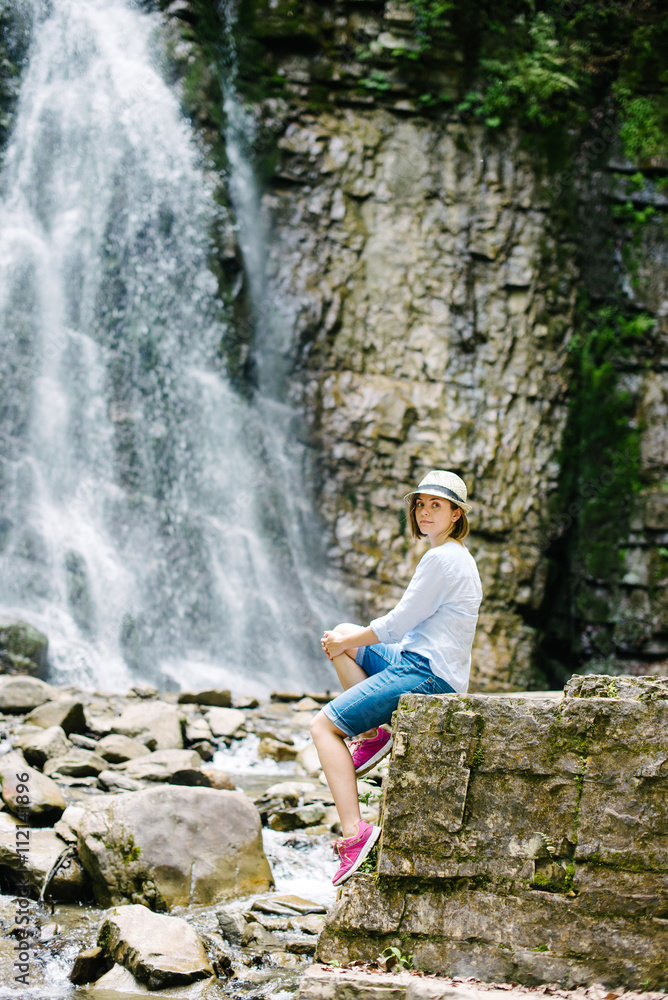 A young girl in a hat sitting on a rock and admires a waterfall in the Carpathian mountains