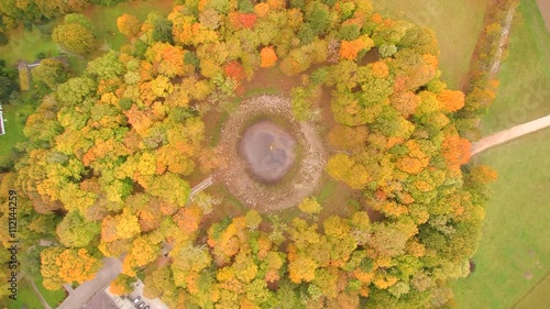 Aerial shot of the beautiful crater with small rocks being surrounded with tall brown-leaf trees in the wide green fields of Kaali in Estonia
