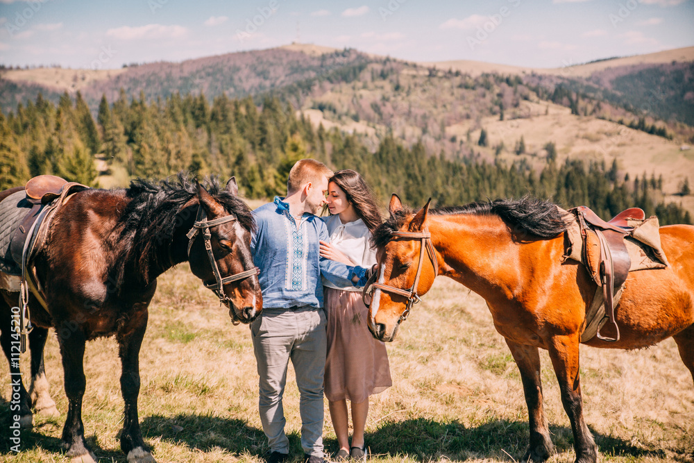 portrait of a cute couple with a horse in a mountains with blues sky. Couple walk and kiss near two horses. portrait of a cute couple with a horse in a mountains with blues sky. Couple walk and kiss near two horses.