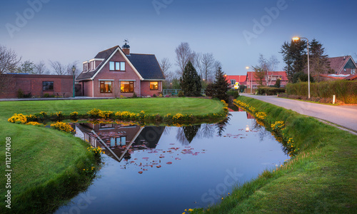 Landscape in dutch village with beautiful house reflected in water canal, courtyard with green grass , yellow flowers and road with illumination at dusk in Netherlands. photo