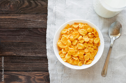 Tasty corn flakes in bowl with bottle of milk. Rustic wooden background with homespun napkin. Healthy crispy breakfast snack. Place for text. Top view, flat lay.