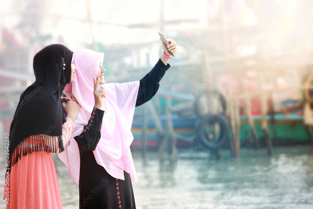 Obraz premium Two happy young muslim girls with Hijab making selfie photo on smartphone.Scene of morning with the mist and soft focus,fishing village