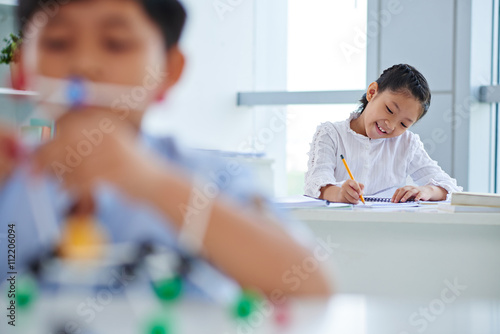 Schoolgirl writing essay in class, her classmate in foreground