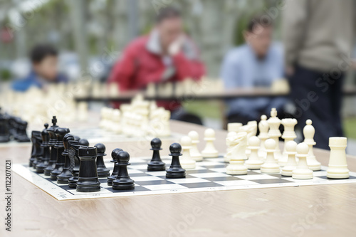 In the foreground a chessboard, in the background (out of focus) several players in a chess simultaneous exhibition photo