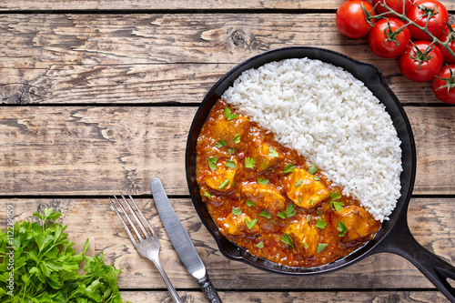 Chicken tikka masala Asian traditional spicy meat food and rice in cast iron skillet with tomatoes, butter, knife. fork and parsley on vintage wooden background. Karahi chicken or korma vindaloo