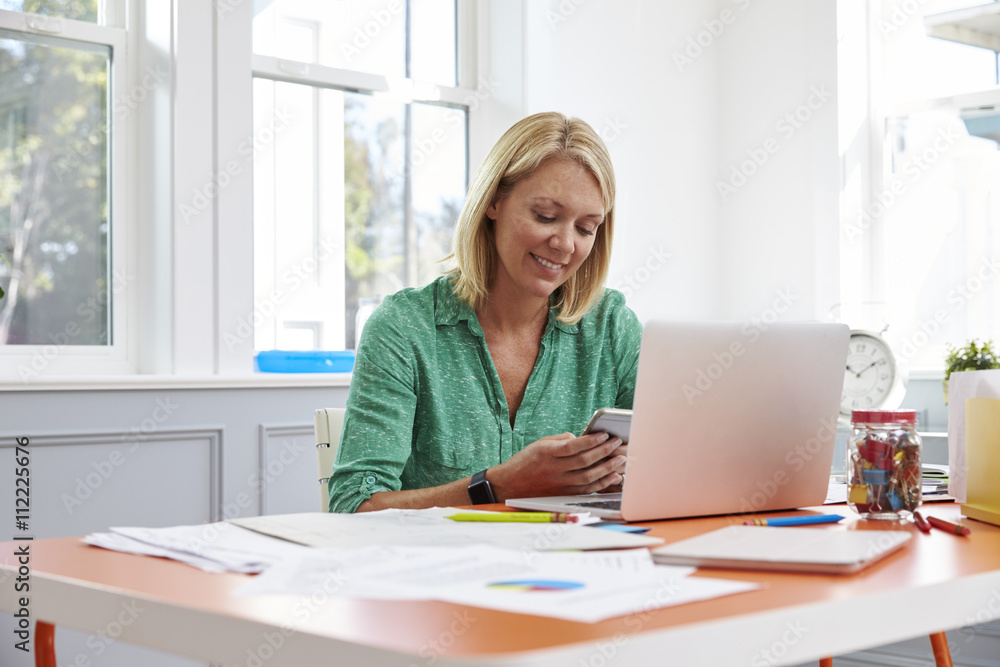 Woman Sitting At Desk Using Mobile Phone In Home Office Woman Sitting At Desk Using Mobile Phone In Home Office
