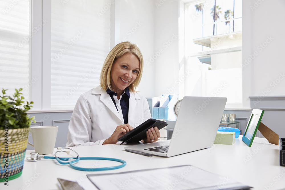 Female Doctor Sitting At Desk Using Digital Tablet In Office Female Doctor Sitting At Desk Using Digital Tablet In Office