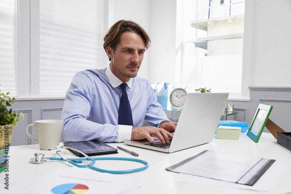 Male Doctor Sitting At Desk Working At Laptop In Office Male Doctor Sitting At Desk Working At Laptop In Office