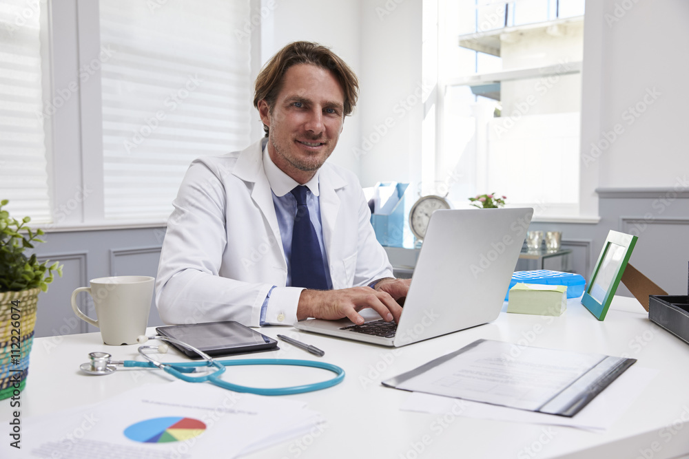 Male Doctor Sitting At Desk Working At Laptop In Office Male Doctor Sitting At Desk Working At Laptop In Office