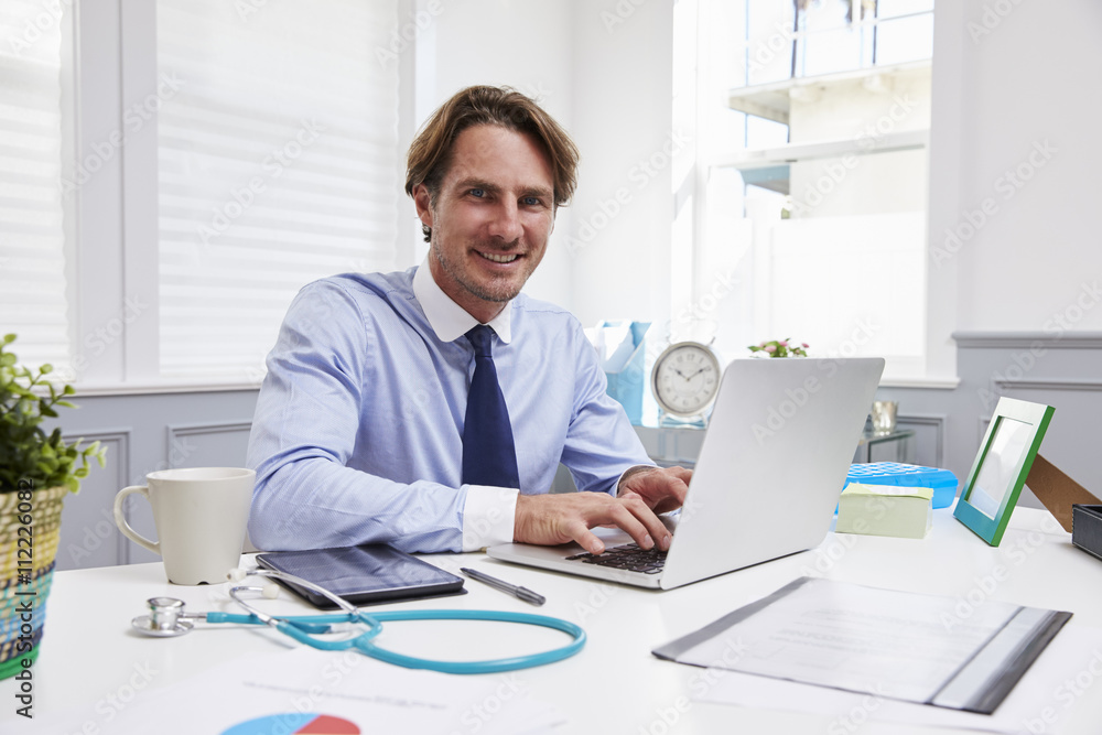 Male Doctor Sitting At Desk Working At Laptop In Office