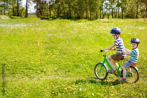 two boys riding on a bicycle. elder brother carries his younger brother on the bike on a blossoming meadow. copy space for your text
