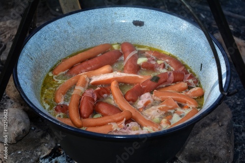 Soup with sausages in iron kettle over campfire 