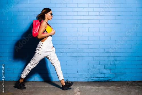 Beautiful young sexy hipster girl goes near urban blue wall background in yellow swimsuit, white jumpsuit, sneakers, sunglasses, pink backpack. photo