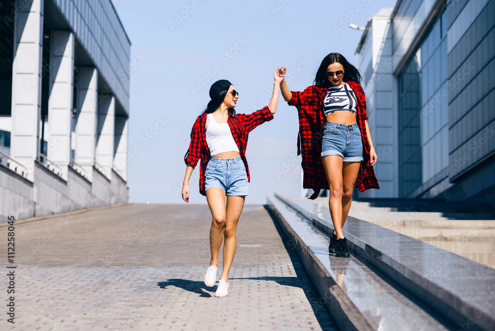 Two young happy hipster girls in sunglasses having fun, smiling, laughing, jumping, walking outdoor on the street, summer relax concept